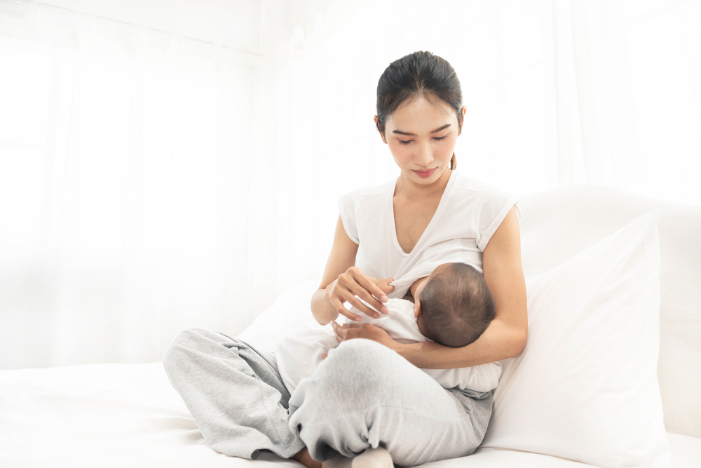 A young woman is sitting on a comfortable bed holding a baby to her breast